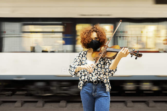 Attractive calm female musician in casual wear and protective face mask playing violin and looking at camera while standing on railway station platform against departing train — Stock Photo