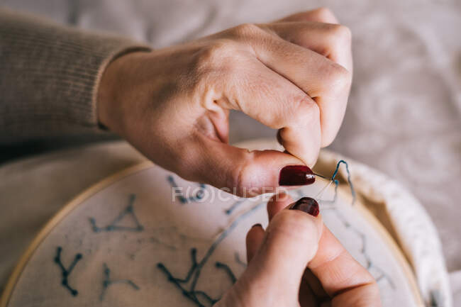 Анонимная женщина с маникюром на руках, нанизывающая иглу, сидя за столом с нитками и обручем и делая вышивку — стоковое фото