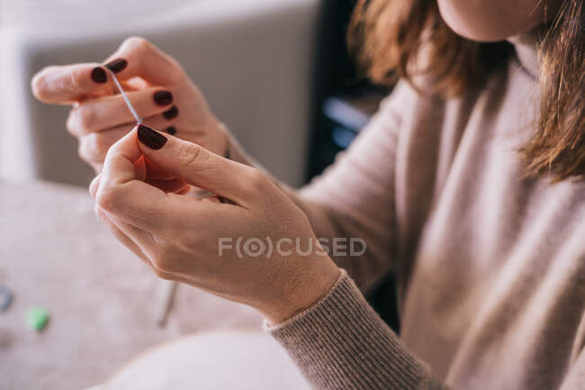 Crop anonymous female in casual outfit threading needle for sewing work while sitting at table in light studio — Stock Photo