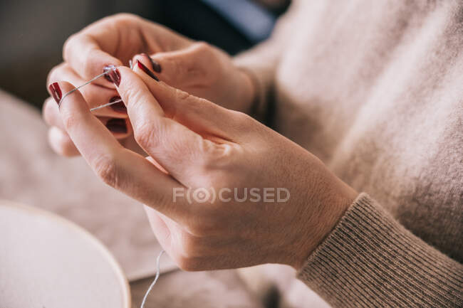 Crop mujer anónima en traje casual que rosca la aguja para el trabajo de costura mientras está sentado en la mesa en el estudio de luz - foto de stock