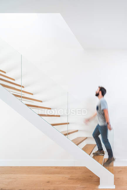 Vue latérale d'un homme flou méconnaissable en tenue décontractée se promenant sur un escalier près d'un mur blanc et regardant vers le haut dans un bâtiment moderne — Photo de stock