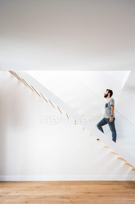 Vista lateral del hombre en traje casual paseando por la escalera cerca de la pared blanca y mirando hacia arriba en el edificio moderno - foto de stock
