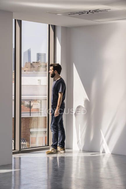Вид сбоку мужчины, стоящего у окна в пустом просторном офисном коридоре с тенями и солнечным светом на белых стенах и смотрящего в сторону — стоковое фото
