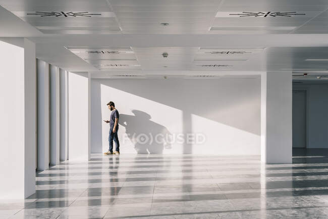 Seitenansicht eines Mannes, der in einem leeren Büroflur mit weißen Wänden und kreativen Schatten steht, während er sein Handy benutzt — Stockfoto