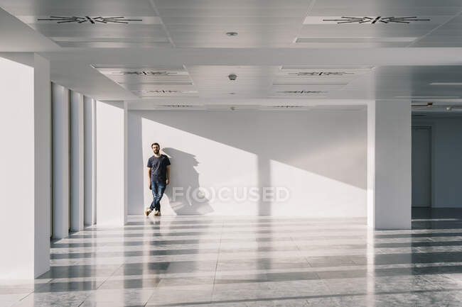 Чоловік стоїть біля вікна в порожньому просторому офісному коридорі з тінями і сонячним світлом на білих стінах і дивиться на камеру — стокове фото