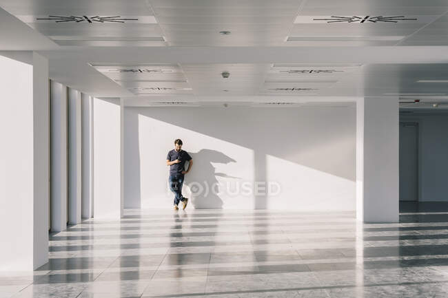 Männchen steht in leerem Büroflur mit weißen Wänden und kreativen Schatten, während sie ihr Handy benutzt — Stockfoto