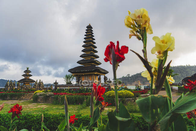 Envejecidos santuarios orientales contra monturas y prados con flores florecientes de colores bajo el cielo nublado en la isla de Bali - foto de stock