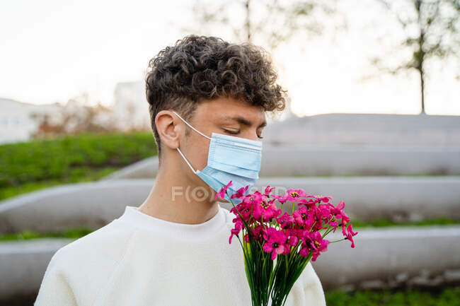 Hombre joven anónimo en máscara estéril con ramo de flores en flor de pie con los ojos cerrados en la ciudad durante la pandemia de COVID 19 - foto de stock