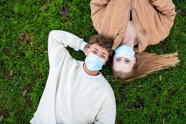 Vista dall'alto di giovani coppie anonime in maschere respiratorie sdraiate sul prato mentre guardano la fotocamera durante la pandemia del COVID 19 — Foto stock