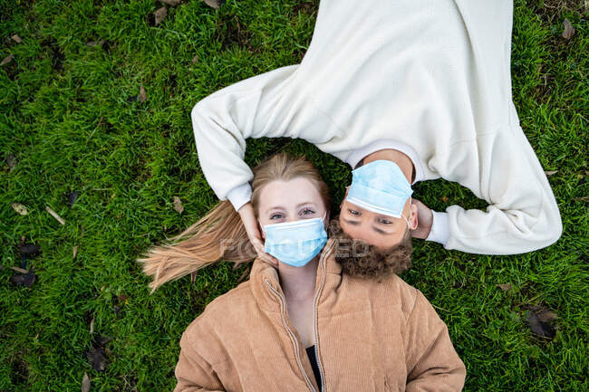 Vista superior de una joven pareja anónima con máscaras respiratorias tumbada en el prado mientras mira la cámara durante la pandemia de COVID 19 - foto de stock