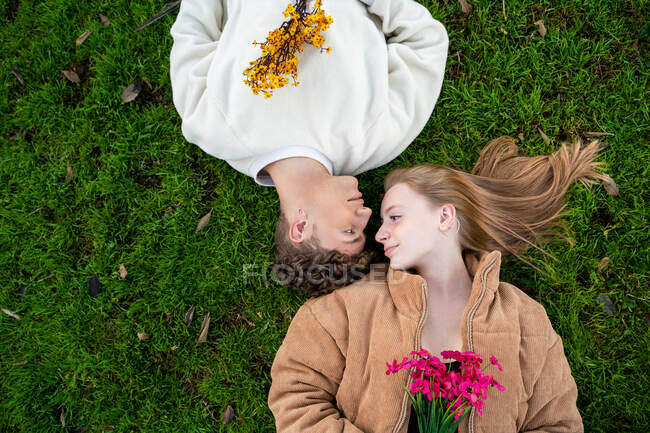 Вид сверху на молодую пару, смотрящую друг на друга, отдыхающую на траве с цветущими букетами цветов — стоковое фото