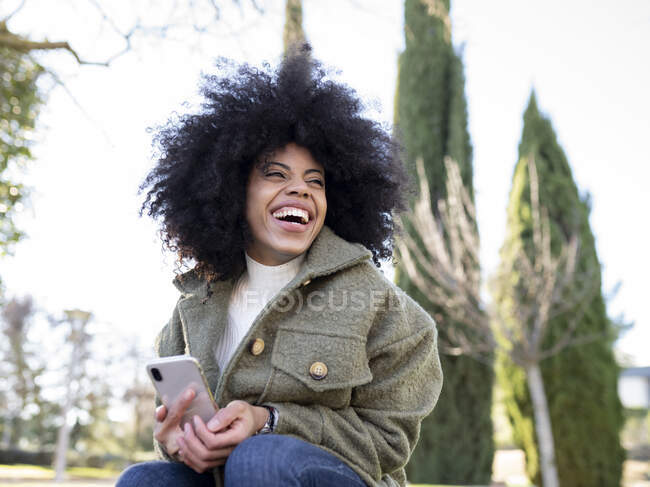 Низкий угол харизматичной молодой афро-американской женщины тысячелетия с кудрявыми волосами смеется и смотрит в сторону во время использования смартфона в парке в солнечный день — стоковое фото