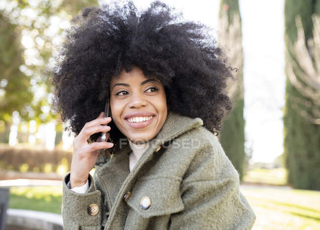 Basso angolo di carismatica giovane afroamericana millenaria con capelli ricci che ride e distoglie lo sguardo mentre usa lo smartphone nel parco nella giornata di sole — Foto stock