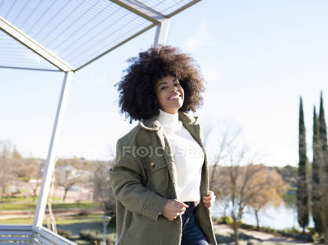 Cheerful young African American female with curly hair in stylish warm outfit smiling and looking at camera while resting in park at lakeside on sunny autumn day — Stock Photo
