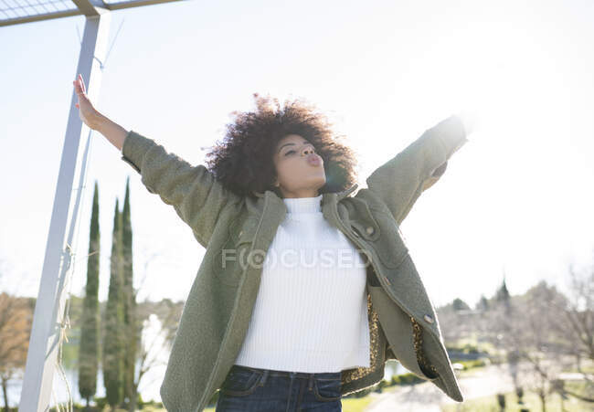 Joven y alegre hembra afroamericana con el pelo rizado en un elegante atuendo cálido con los brazos abiertos mientras descansa en el parque junto al lago en el soleado día de otoño. - foto de stock