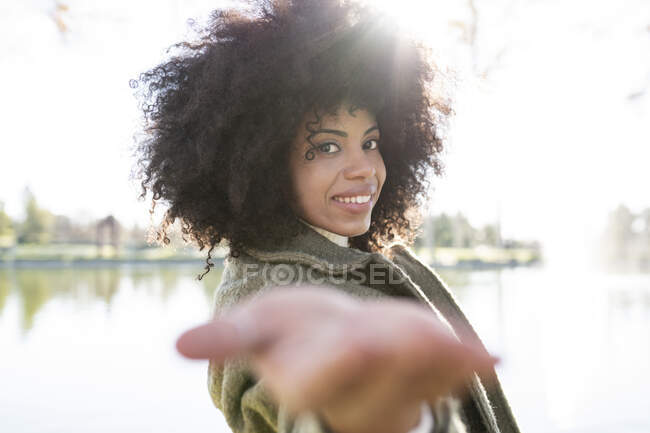 Вид сбоку на позитивную молодую черную леди с прической афро в модном наряде протягивающую руку к камере и улыбающуюся во время отдыха у озера в осеннем парке — стоковое фото
