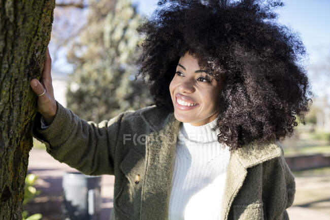 Fröhliche junge Afroamerikanerin mit lockigem Haar in stylischem warmem Outfit lächelt und schaut weg, während sie sich an sonnigen Herbsttagen im Park am Seeufer ausruht — Stockfoto