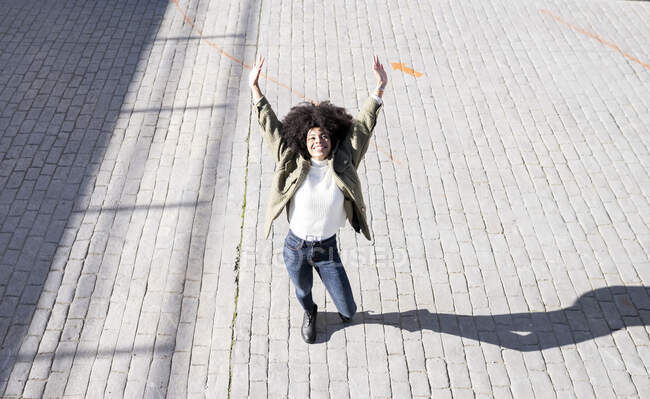 De cima de expressiva jovem senhora negra com penteado afro em roupas elegantes de pé na rua paves com braços levantados e sorrindo no dia ensolarado — Fotografia de Stock