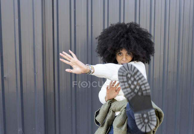 Auto assuré jeune dame ethnique avec coiffure afro en tenue à la mode et bottes bottant caméra tout en se tenant debout sur la rue près du mur de métal — Photo de stock