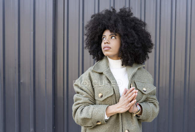 Pensive young stylish African American female millennial with curly hair in trendy outfit standing on street near metal fence and looking away — Stock Photo