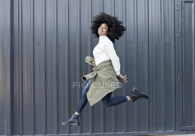 Вид сбоку на счастливую молодую афроамериканку с вьющимися волосами в модном наряде, улыбающуюся и смотрящую в камеру во время прыжка на улице — стоковое фото