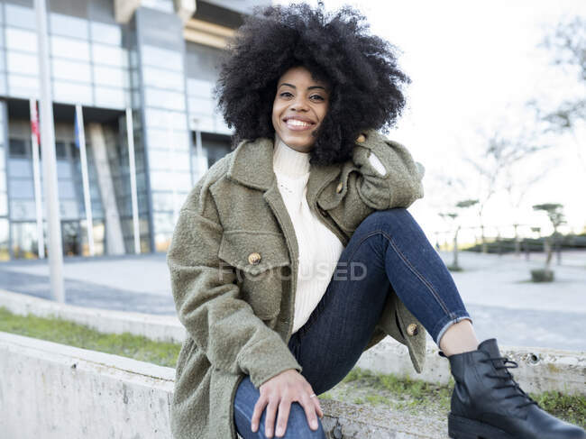 Moda joven negro femenino millennial con pelo afro en ropa de abrigo con estilo descansando en la calle y mirando a la cámara pensativamente cerca de un edificio moderno con paredes de vidrio - foto de stock