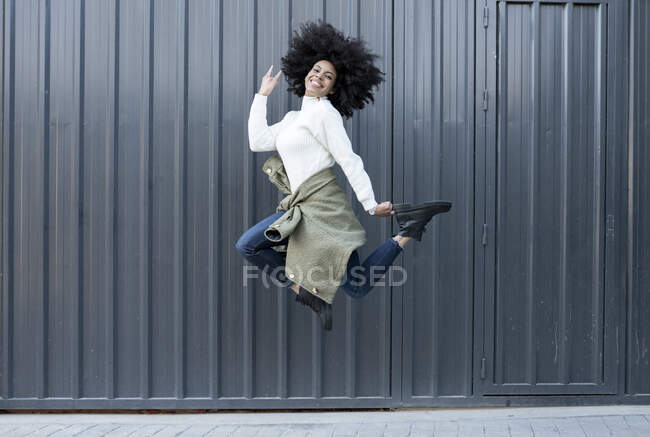 Vista lateral de la joven mujer afroamericana feliz con el pelo rizado en traje de moda sonriendo y mirando a la cámara mientras salta a la calle - foto de stock