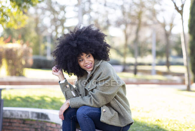 Moda joven negro femenino millennial con pelo afro en ropa de abrigo con estilo descansando en la calle y mirando hacia otro lado pensativamente - foto de stock