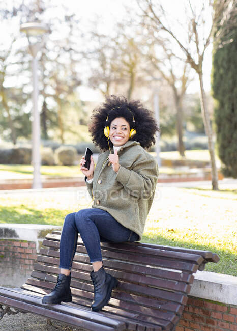 Full body of excited young African American female millennial with curly hair in stylish outfit resting on bench in park and listening favorite song in headphones — Stock Photo