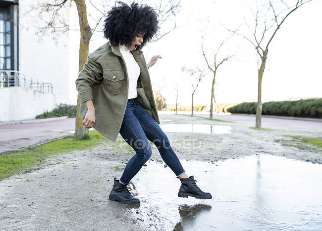 Vue latérale de la jeune femme afro-américaine expressive millénium en tenue tendance trempant le pied dans l'eau de la flaque d'eau tout en se tenant dans la rue par une journée ensoleillée — Photo de stock