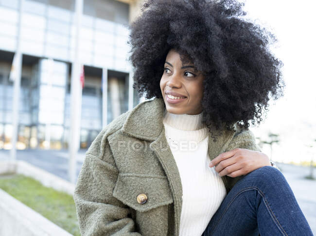 Tendance jeune millénium féminin noir avec des cheveux afro dans des vêtements chauds élégants reposant sur la rue et regardant loin pensivement près du bâtiment moderne avec des murs en verre — Photo de stock