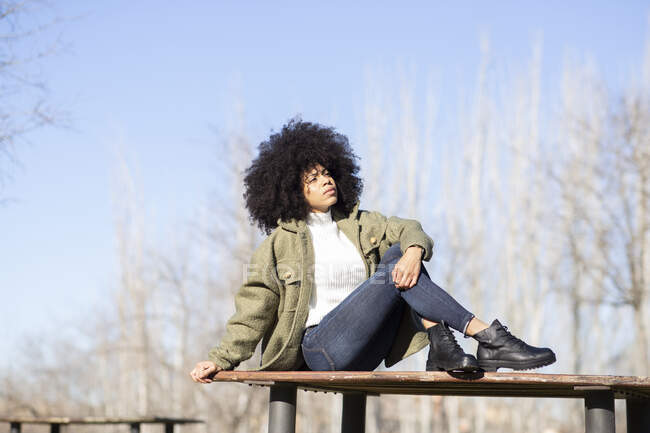 Cuerpo completo de mujer afroamericana joven reflexiva con cabello rizado oscuro en ropa de abrigo de moda relajándose en el banco de madera en el parque y mirando hacia otro lado soñando en el soleado día de otoño - foto de stock
