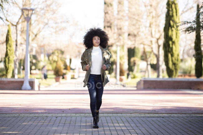 Повний набір трендів молода афроамериканська леді з кучерявим волоссям в повсякденному вбранні ходячи в міському парку і задумливо озираючись геть — стокове фото