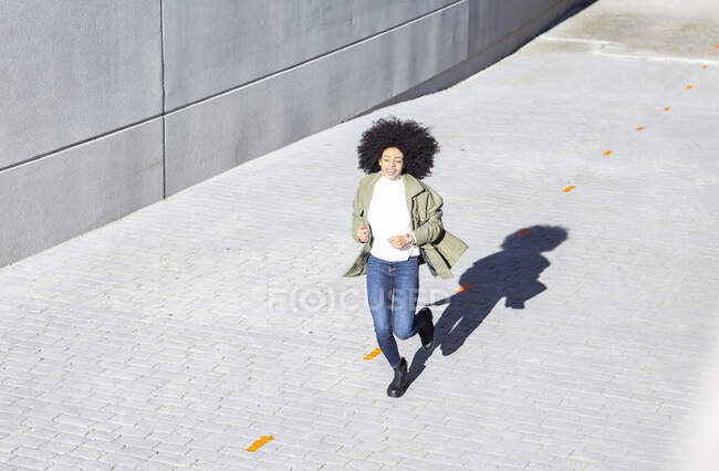 Desde arriba de la joven expresiva negra con peinado afro en ropa elegante caminando por la calle pavimentos y sonriendo en el día soleado - foto de stock