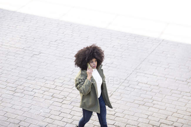High angle of concentrated young African American female millennial with curly hair in stylish outfit having phone conversation while walking on paved street on sunny day — Stock Photo