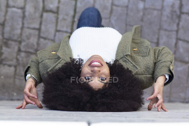 Вид сверху на молодую афроамериканку с кудрявыми волосами в стильной одежде, опирающуюся на стену и смотрящую вверх, стоя на мощеной улице в солнечный день — стоковое фото