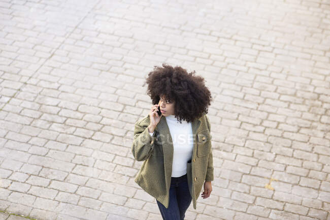 Високий кут зосередженої молодої афроамериканської жінки тисячоліття з кучерявим волоссям в стильному одязі, який розмовляє по телефону під час ходьби по брукованій вулиці в сонячний день. — стокове фото