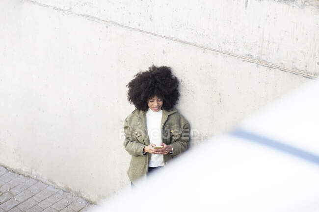 Angle élevé de charismatique jeune afro-américaine millénaire avec les cheveux bouclés rire et détourner les yeux tout en utilisant smartphone dans le parc par jour ensoleillé — Photo de stock