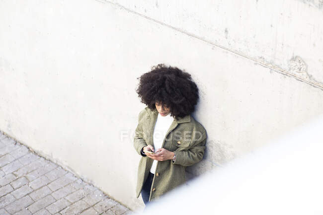 Високий кут харизматичної молодої афроамериканської жінки тисячоліття з кучерявим волоссям при використанні смартфона на вулиці в сонячний день. — стокове фото