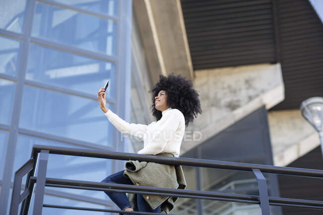 Vista lateral de la joven mujer negra confiada con peinado afro en ropa casual sonriendo mientras toma selfie en el teléfono inteligente de pie en el balcón del edificio moderno - foto de stock