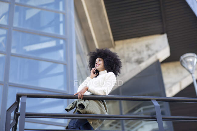 Vista lateral da jovem mulher negra confiante com penteado afro em roupas casuais sorrindo enquanto conversa no smartphone em pé na varanda do edifício moderno — Fotografia de Stock