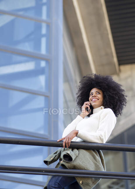 Vue latérale de la jeune femme noire confiante avec coiffure afro dans des vêtements décontractés souriant tout en parlant sur smartphone debout sur le balcon du bâtiment moderne — Photo de stock