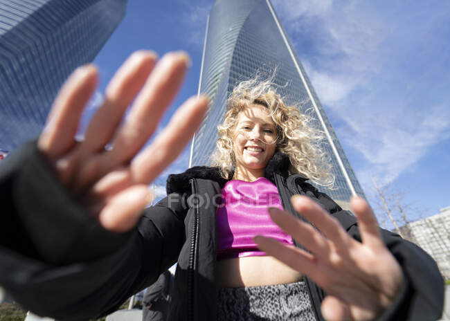 Знизу весела жінка з блондинкою кучеряве волосся і в вишуканому вбранні, що тягнеться до камери, стоячи в центрі міста проти хмарочосів в сонячний день — стокове фото