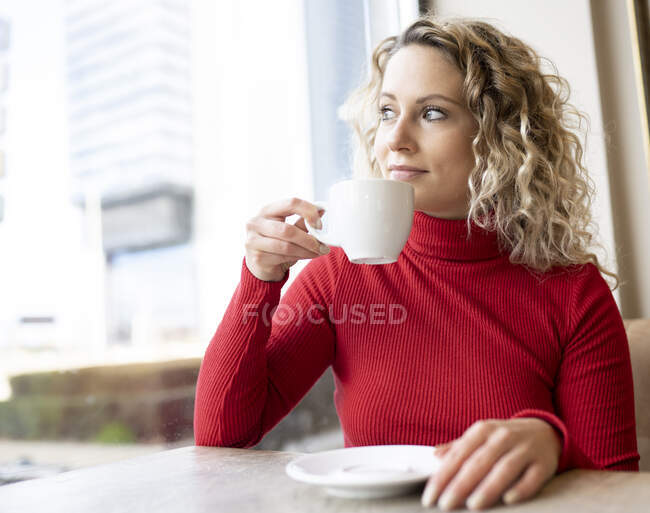 Mujer soñadora sentada en la mesa en la cafetería y bebiendo bebidas calientes mientras disfruta del fin de semana y mira por la ventana - foto de stock