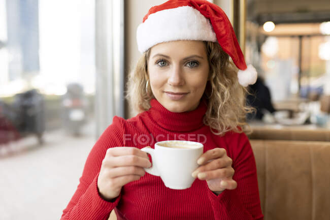 Mujer encantada en Santa sombrero y cuello alto rojo sentado con taza de café en la cafetería y mirando a la cámara durante las vacaciones de Navidad - foto de stock