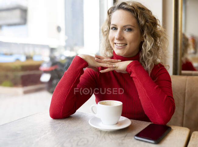 Verträumte Frau sitzt am Tisch im Café und trinkt Heißgetränk, während sie das Wochenende genießt und in die Kamera schaut — Stockfoto