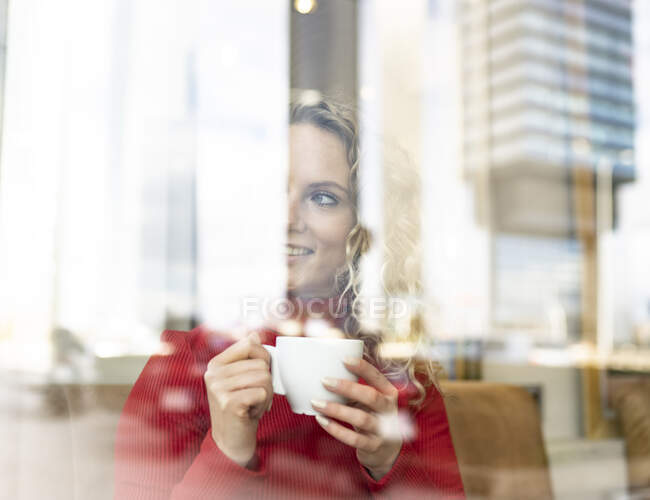 Através da janela de mulher sonhadora sentada à mesa no café e bebendo bebida quente enquanto desfruta de fim de semana e olhando pela janela — Fotografia de Stock