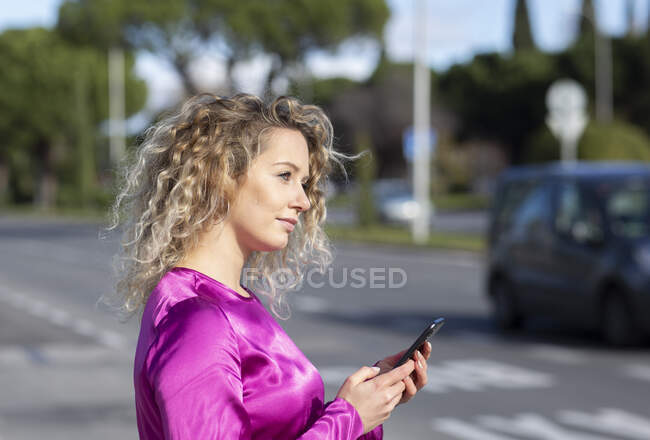Vista lateral de una mujer de ensueño con el pelo rubio rizado de pie cerca de la carretera en la ciudad y el uso de un teléfono inteligente mientras mira hacia otro lado en la contemplación - foto de stock