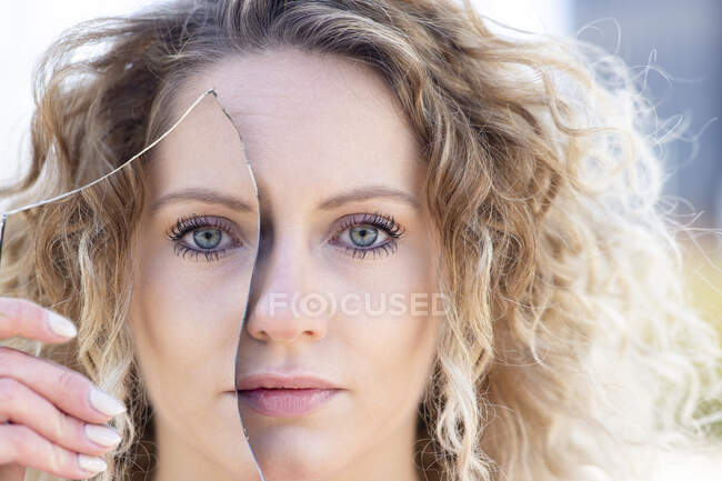 Mujer sin emociones con cabello rubio rizado que cubre el ojo con un trozo de espejo roto con reflejo de la cara y mirando a la cámara - foto de stock