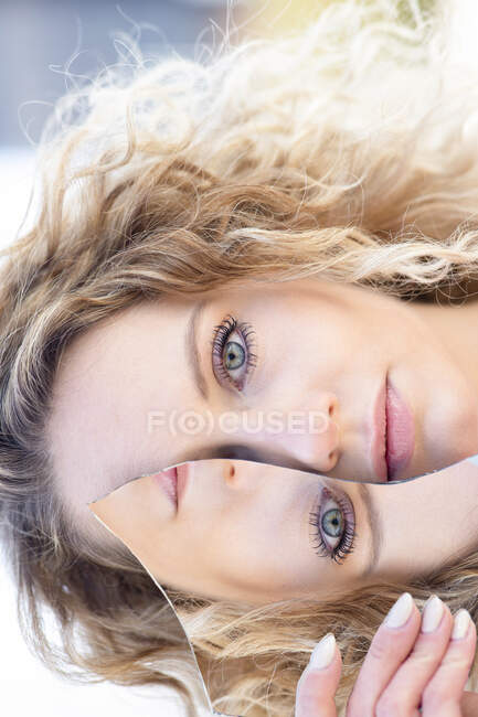 Mujer sin emociones con cabello rubio rizado que cubre el ojo con un trozo de espejo roto con reflejo de la cara y mirando a la cámara - foto de stock